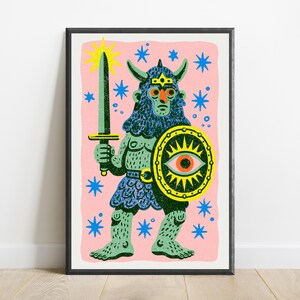 May include: A framed art print featuring a green horned warrior figure with a blue beard and textured skin. The figure holds a green sword and a yellow shield with a central eye motif. The background is light pink with scattered blue stars.