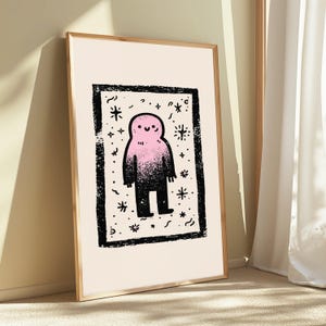 May include: A framed wall art print featuring a central illustration of a smiling, rounded creature with a pink head that fades into a black body. The figure is enclosed within a thick, textured black rectangular border on a cream background.