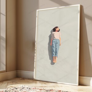 May include: A framed vertical wall art print featuring a minimalist illustration of a person lying down on a light beige background. The person wears a light pink short-sleeved shirt and light blue trousers, casting a dark shadow on the surface.