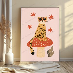 Op de afbeelding: Een ingelijste muurkunstprint met een gele jachtluipaard met zwarte vlekken die een ronde oranje zonnebril draagt, zittend op een grote rode paddenstoel met witte stippen. De achtergrond is lichtroze met verspreide rode sterren en een plant.