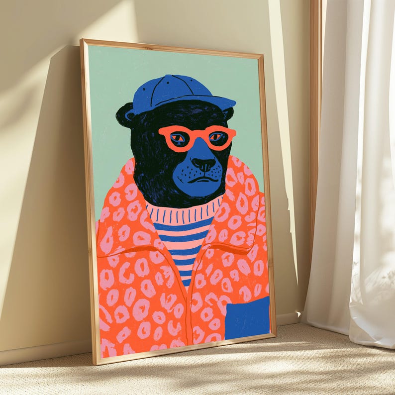May include: A framed art print featuring a black bear wearing a blue baseball cap, bright orange glasses, and a vibrant orange jacket with a pink leopard print pattern over a blue and pink striped shirt, set against a light green background.