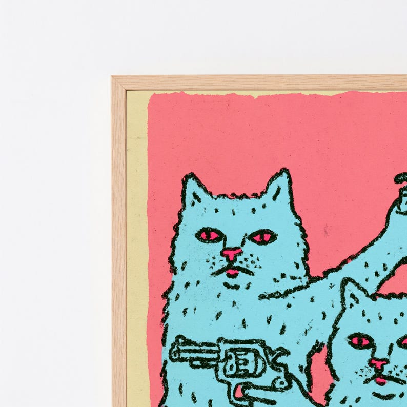 May include: A framed art print featuring two light blue cats with pink eyes against a vibrant pink background. The larger cat holds a black outlined revolver in its paw. The illustration uses a bold, textured black line style throughout the design.