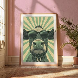 May include: A framed wall art print featuring a stylized illustration of a cow wearing black sunglasses. The cow is centered against a background of alternating light green and cream-colored sunburst rays, creating a bold graphic composition.