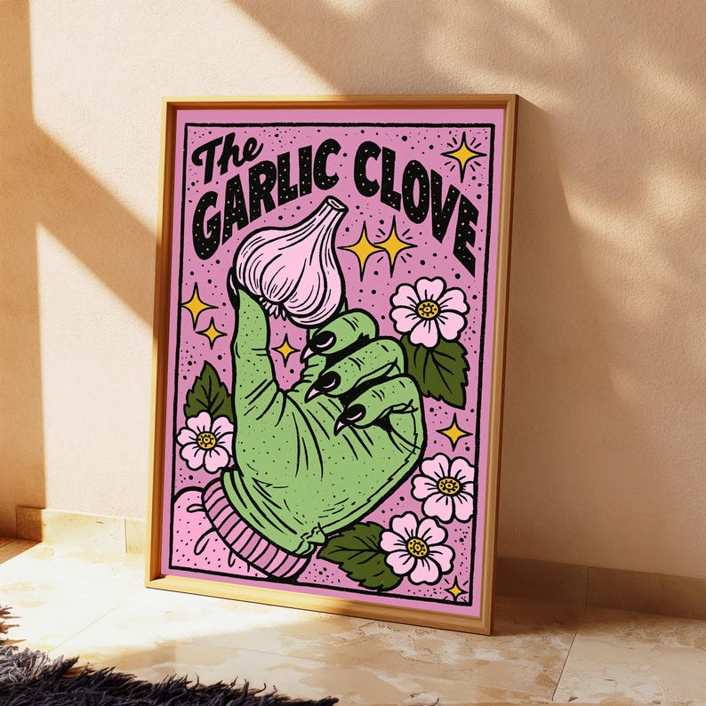May include: A framed rectangular wall art print featuring a green hand with black fingernails holding a pink garlic clove. The background is light pink with black text reading The Garlic Clove, surrounded by small yellow stars and pink flowers.