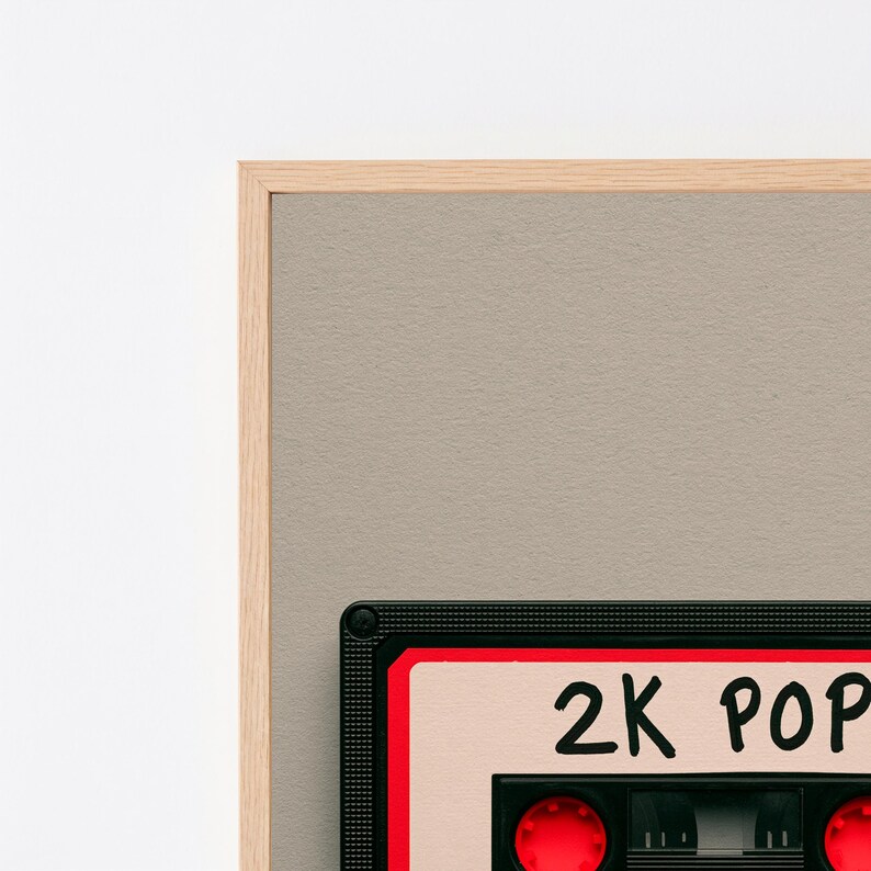May include: A framed wall art piece featuring a graphic illustration of a black cassette tape with red accents and a beige label. The label displays the text 2K POP in black handwritten-style lettering. The frame is made of light-coloured wood.