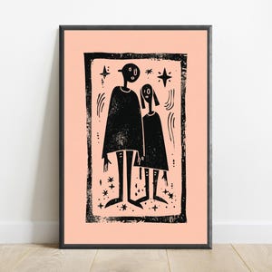 May include: A framed wall art print featuring a black ink block print illustration of two stylized figures standing side by side on a light peach background. The artwork includes small star shapes and curved lines, all enclosed within a textured black border.