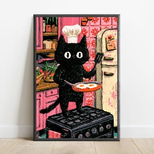 Peut inclure: Une impression d'art encadr&eacute;e repr&eacute;sentant un chat noir portant une toque de chef blanche, debout sur une cuisini&egrave;re noire dans une cuisine rose. Le chat tient une spatule noire et une po&ecirc;le rouge contenant deux &oelig;ufs au plat, devant des meubles roses.