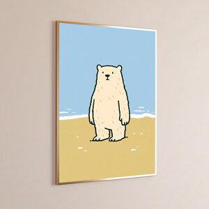May include: A framed wall art print featuring a cartoon illustration of a cream-colored bear standing on a tan sandy beach. The background shows a light blue sky and a thin white line representing the ocean waves meeting the shore.