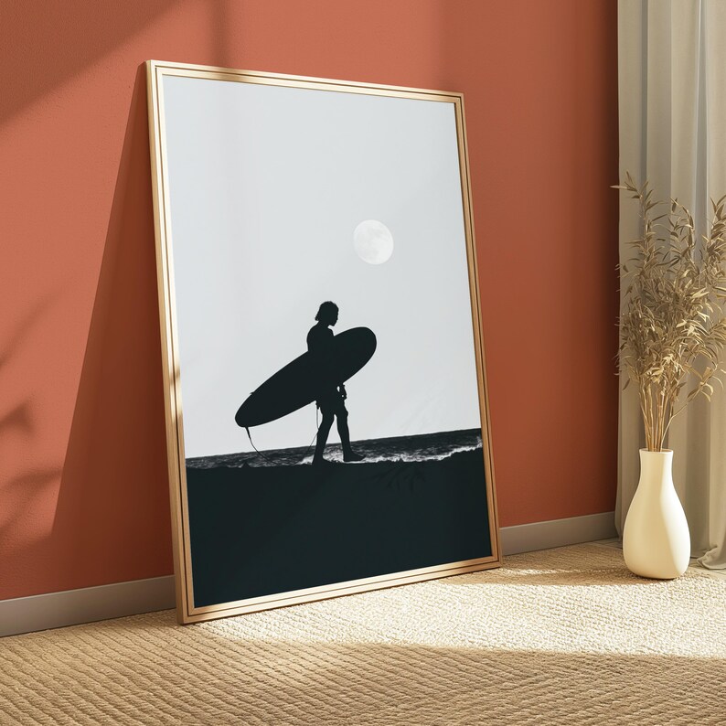 May include: A framed vertical wall art print featuring a black silhouette of a person carrying a surfboard along a dark shoreline against a light gray sky with a pale moon. The artwork is enclosed in a thin, light-colored wooden frame.