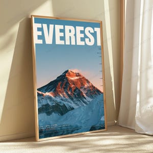 Op de afbeelding: Een ingelijste verticale poster met een grafische illustratie van de Mount Everest. Het woord EVEREST staat bovenaan in grote witte letters gedrukt. De bergtop wordt verlicht door oranje zonlicht tegen een blauwe lucht en besneeuwde hellingen.
