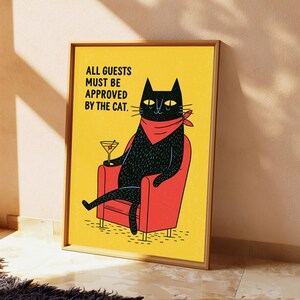 May include: A framed wall art print featuring a black cat wearing a red bandana, sitting in a red armchair, holding a martini glass. The yellow background includes the black text: ALL GUESTS MUST BE APPROVED BY THE CAT. The print is in a wooden frame.
