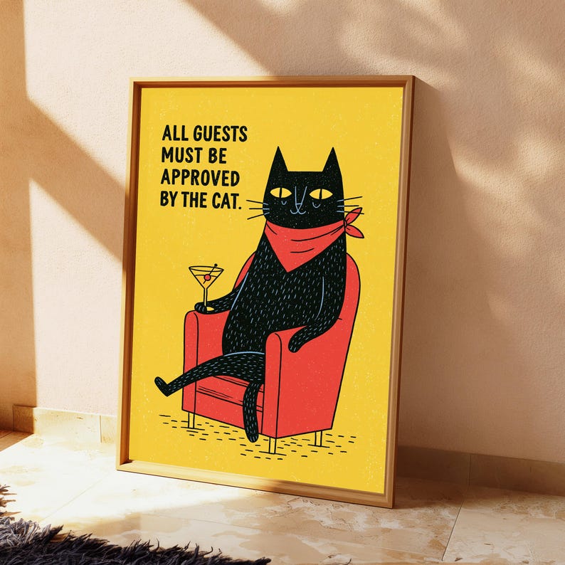 May include: A framed wall art print featuring a black cat wearing a red bandana, sitting in a red armchair, holding a martini glass. The yellow background includes the black text: ALL GUESTS MUST BE APPROVED BY THE CAT. The print is in a wooden frame.