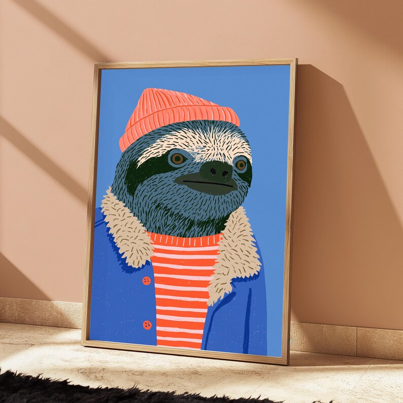 May include: A framed art print featuring a stylized illustration of a sloth wearing a coral-colored beanie, a red and white striped shirt, and a blue jacket with a beige faux-fur collar, all set against a solid light blue background.