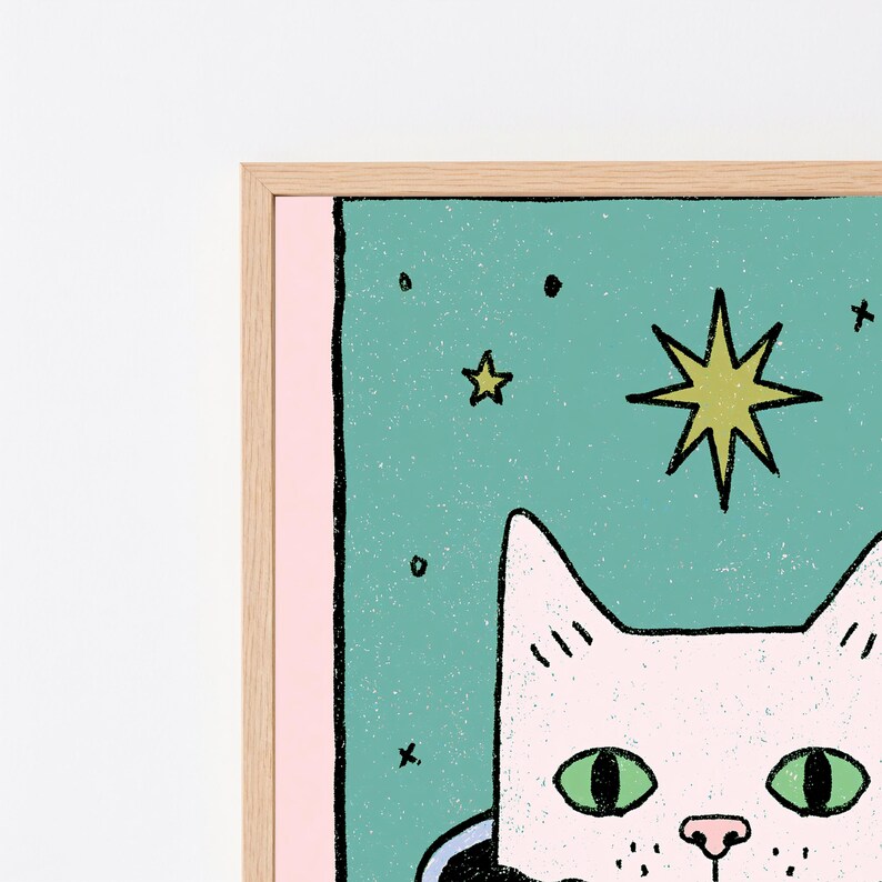 May include: A framed wall art print featuring a light pink cat with green eyes centered against a teal background. The illustration includes a large yellow star, smaller black stars, and a light pink border, all enclosed in a light wood frame.