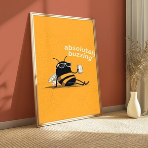 May include: A framed rectangular wall art print featuring a yellow background with a cartoon bee wearing white sunglasses and holding a silver can. The text "absolutely buzzing" is printed in white above the bee, which has black and yellow stripes.