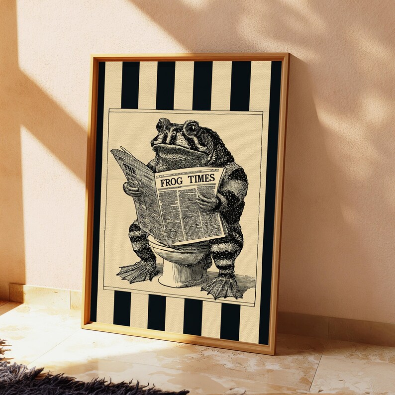 May include: A framed wall art print featuring a black and white illustration of a toad sitting on a toilet reading a newspaper titled FROG TIMES. The central image is set against a background of vertical black and beige stripes within a wooden frame.
