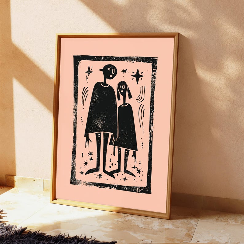 May include: A framed wall art print featuring a black ink block print illustration of two stylized figures standing side by side. The figures are surrounded by small star shapes and curved lines, all enclosed within a textured black rectangular border on a solid light peach background.