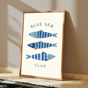 Pode incluir: Uma impress&atilde;o de arte emoldurada apresentando tr&ecirc;s peixes orientados horizontalmente com listras azuis e brancas. O texto BLUE SEA est&aacute; impresso em azul acima dos peixes e a palavra CLUB est&aacute; impressa em azul abaixo deles em um fundo creme.