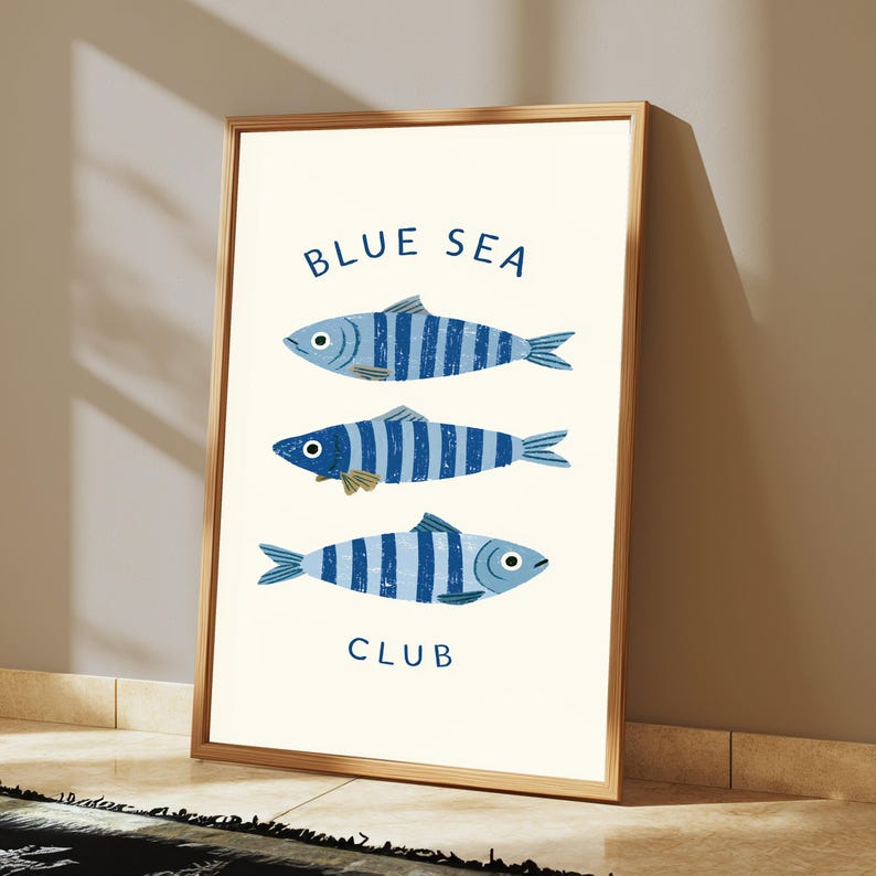 Pode incluir: Uma impress&atilde;o de arte emoldurada apresentando tr&ecirc;s peixes orientados horizontalmente com listras azuis e brancas. O texto BLUE SEA est&aacute; impresso em azul acima dos peixes e a palavra CLUB est&aacute; impressa em azul abaixo deles em um fundo creme.
