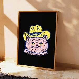 Puede incluir: Una l&aacute;mina de arte enmarcada que presenta una ilustraci&oacute;n estilizada de un gato rosa con un sombrero de vaquero amarillo sobre un fondo negro s&oacute;lido. El gato tiene ojos amarillos y contornos azul oscuro, en un marco de madera de color claro.