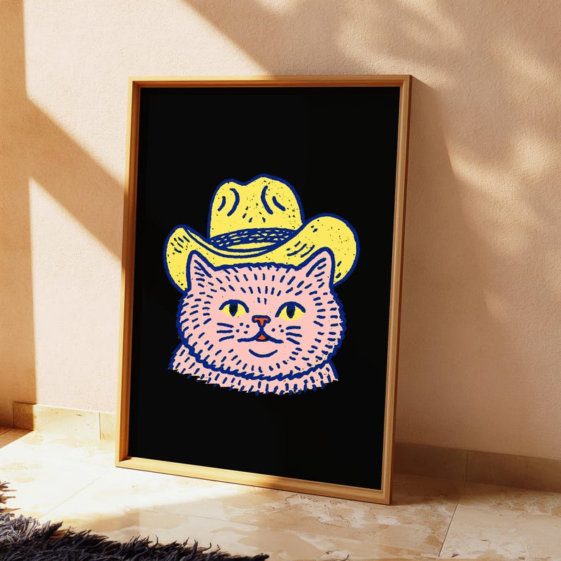 Puede incluir: Una l&aacute;mina de arte enmarcada que presenta una ilustraci&oacute;n estilizada de un gato rosa con un sombrero de vaquero amarillo sobre un fondo negro s&oacute;lido. El gato tiene ojos amarillos y contornos azul oscuro, en un marco de madera de color claro.