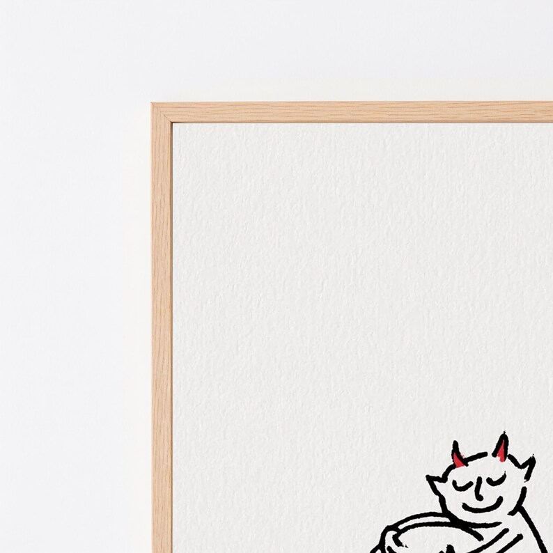 May include: A framed wall art piece featuring a minimalist black line drawing of a small creature with red horns, shown with eyes closed and a gentle smile, holding a round object against a plain white background within a light wood frame.