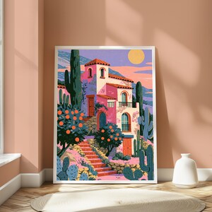 May include: A framed art print depicting a pink Mediterranean-style villa with arched windows and balconies. The scene features orange trees, tall green cacti, and a stone staircase leading to the house under a pale yellow sun in a pastel sky.