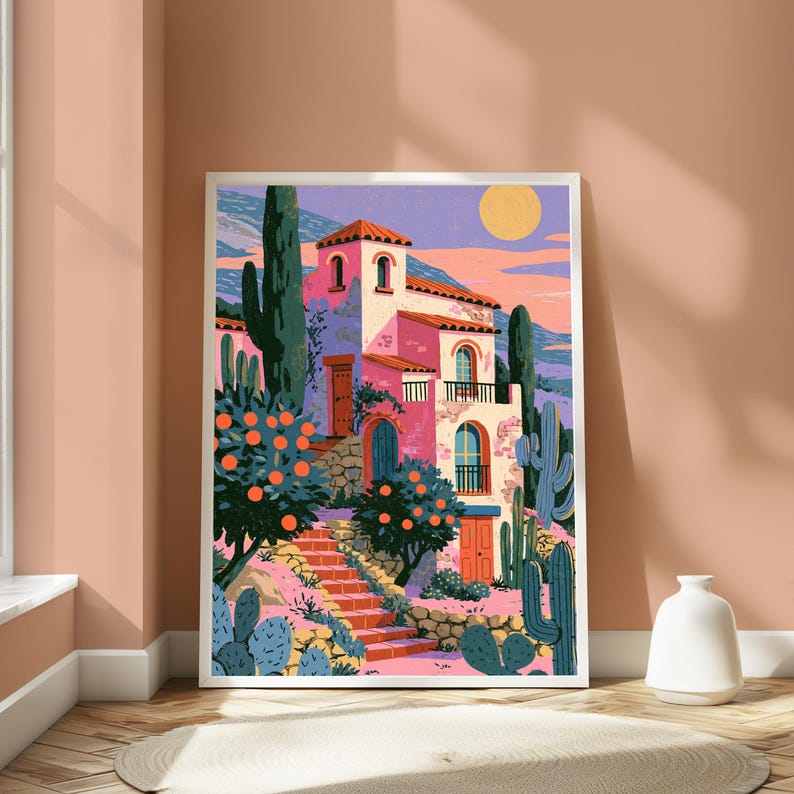 May include: A framed art print depicting a pink Mediterranean-style villa with arched windows and balconies. The scene features orange trees, tall green cacti, and a stone staircase leading to the house under a pale yellow sun in a pastel sky.