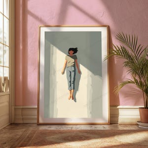 May include: A framed wall art print featuring a stylized illustration of a person with dark hair, wearing a beige t-shirt and blue cropped pants, floating in a vertical beam of light against a muted sage green background with sharp shadows.
