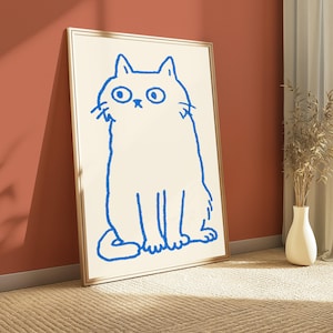 May include: A framed wall art print featuring a simple, hand-drawn style illustration of a cat in bright blue ink on a cream-colored background. The cat is depicted in a seated position with large, wide eyes and a neutral expression.