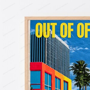 May include: A framed wall art print featuring a tall skyscraper against a blue sky with palm trees. The text "OUT OF OF" is printed in large yellow letters at the top. The building base is composed of red, orange, and yellow rectangular sections.