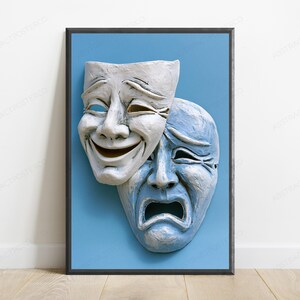 May include: A framed wall art print featuring two theatrical masks against a solid light blue background. The top mask is white and displays a smiling expression, while the partially overlapping bottom mask is light blue and displays a sad expression.