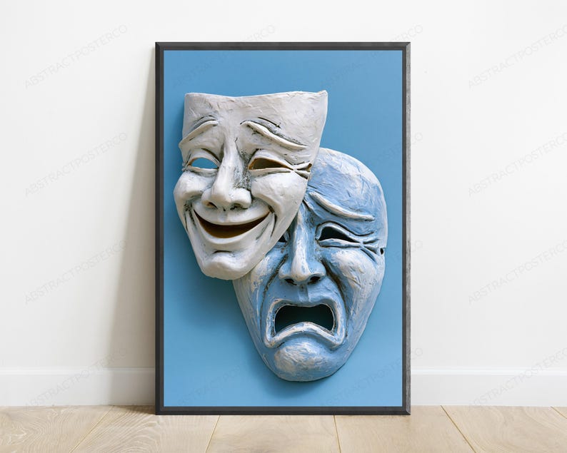 May include: A framed wall art print featuring two theatrical masks against a solid light blue background. The top mask is white and displays a smiling expression, while the partially overlapping bottom mask is light blue and displays a sad expression.