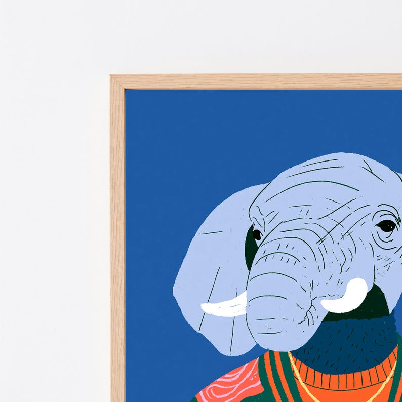 May include: A framed art print featuring a light blue elephant head against a solid blue background. The elephant wears a dark green and orange sweater with a ribbed collar. The frame is made of light-colored wood with a simple, clean profile.