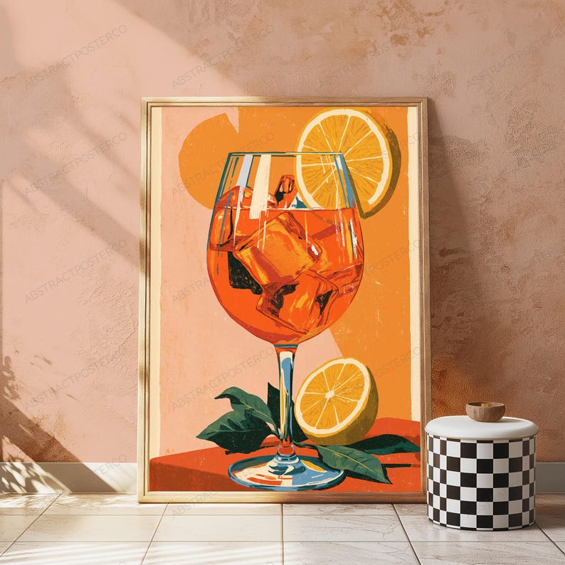 May include: A framed art print featuring a vibrant orange cocktail in a stemmed glass filled with ice cubes. An orange slice garnish rests on the rim, and another orange slice sits at the base with green leaves against a warm orange background.