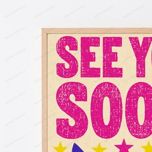 May include: A framed wall art print featuring the bright pink text SEE YOU SOON in a distressed, textured font against a light beige background. Below the text, a row of small, colorful stars in yellow, blue, and pink are visible in the frame.