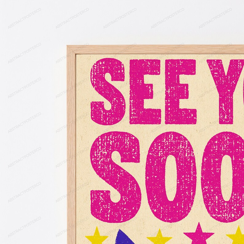 May include: A framed wall art print featuring the bright pink text SEE YOU SOON in a distressed, textured font against a light beige background. Below the text, a row of small, colorful stars in yellow, blue, and pink are visible in the frame.