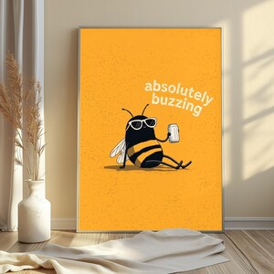 May include: A rectangular wall art print featuring a cartoon bee with black and yellow stripes, wearing white-rimmed sunglasses, sitting and holding a silver can. The background is solid orange with the white text absolutely buzzing.