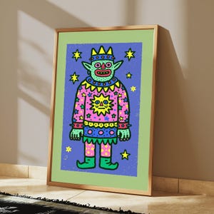 May include: A framed art print featuring a cartoon creature with green skin, a crown, and a pink patterned tunic decorated with a yellow sun and stars. The figure stands against a blue background filled with yellow stars, all inside a light green border.