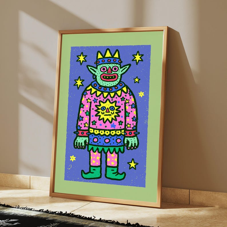 May include: A framed art print featuring a cartoon creature with green skin, a crown, and a pink patterned tunic decorated with a yellow sun and stars. The figure stands against a blue background filled with yellow stars, all inside a light green border.