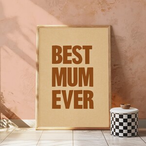 May include: A framed print with the text "BEST MUM EVER" in brown capital letters. The print has a light tan background and a gold-colored frame. A black and white checkered container with a wooden lid is positioned to the right.