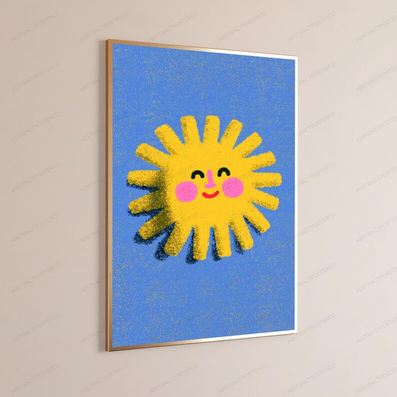 May include: A rectangular wall art print in a thin gold frame featuring a textured yellow sun illustration on a blue background. The sun has a smiling face with black eyes, a small pink nose, pink cheeks, and a red curved mouth, all with a grainy texture.