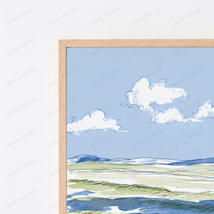 May include: A framed wall art print featuring a landscape illustration with a light blue sky, white clouds, and rolling green and blue fields. The artwork is enclosed in a light-colored wooden frame with a visible grain texture and clean edges.