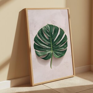 May include: A framed wall art print featuring a single, large, dark green monstera leaf centered on a pale, textured, light pink background. The artwork is enclosed in a simple, light-toned wooden frame with a clean, rectangular profile.