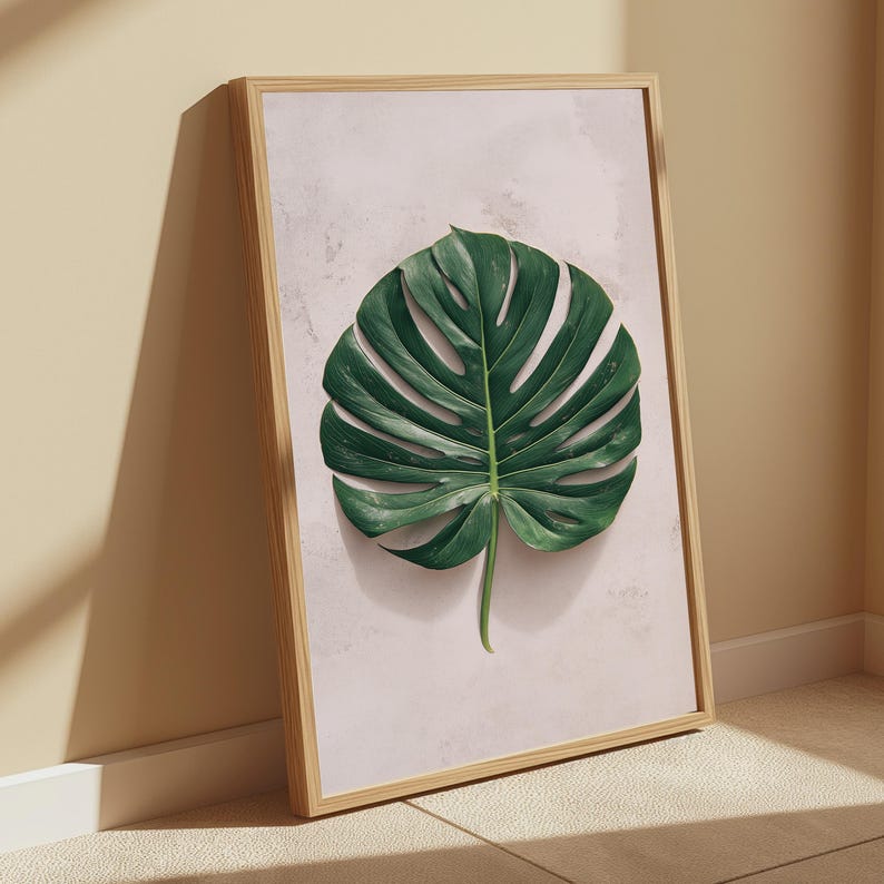 May include: A framed wall art print featuring a single, large, dark green monstera leaf centered on a pale, textured, light pink background. The artwork is enclosed in a simple, light-toned wooden frame with a clean, rectangular profile.