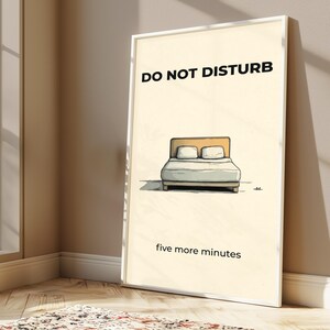 Puede incluir: Un cuadro enmarcado con las palabras "DO NOT DISTURB" sobre una ilustraci&oacute;n de una cama. Debajo de la cama, el texto dice "five more minutes". La obra de arte tiene un estilo minimalista con un fondo crema.
