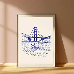 May include: A framed wall art print featuring a blue ink line drawing on a cream background. The illustration depicts a suspension bridge spanning a body of water with a small boat and a person fishing in the foreground, set against hilly terrain.