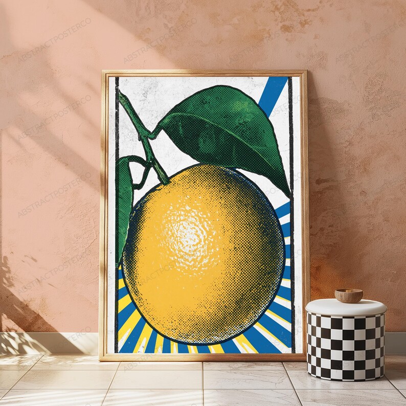 May include: A framed wall art print featuring a stylized yellow orange with green leaves. The artwork uses a halftone dot pattern and is set against a background of radiating blue and white sunburst lines, enclosed within a black rectangular border.