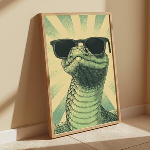 May include: A framed wall art print featuring a green snake wearing black sunglasses. The illustration shows the snake from the chest up against a background of light yellow and teal sunburst rays. The artwork is encased in a light wood frame.