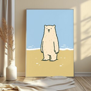 May include: A framed wall art print featuring a cartoon illustration of a cream-colored polar bear standing on a sandy yellow beach. The background shows a light blue sky and a thin white line representing the ocean waves in a simple style.